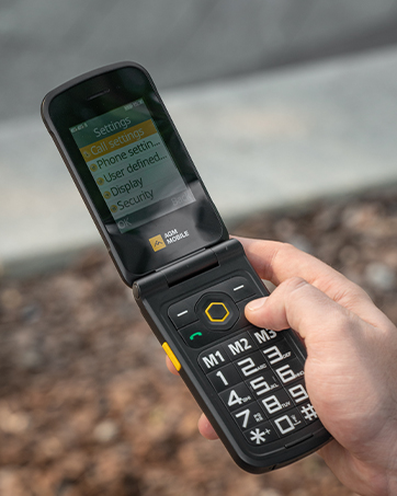 Rugged black feature phone being held in hand, showing keypad and small display screen, with building facade visible in background.