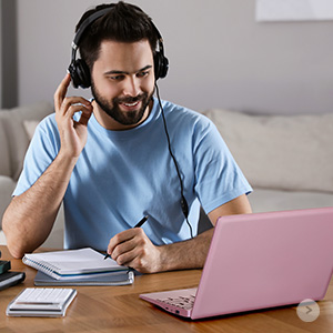 Man wearing headphones while working on a laptop. He's writing in a notebook, suggesting a remote work or study setup.