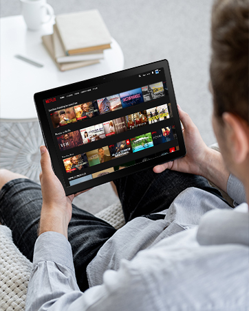 Tablet device held in hands displaying video conference with six participants on screen. Person wearing blue shirt sits at desk with coffee cup and desk accessories visible.