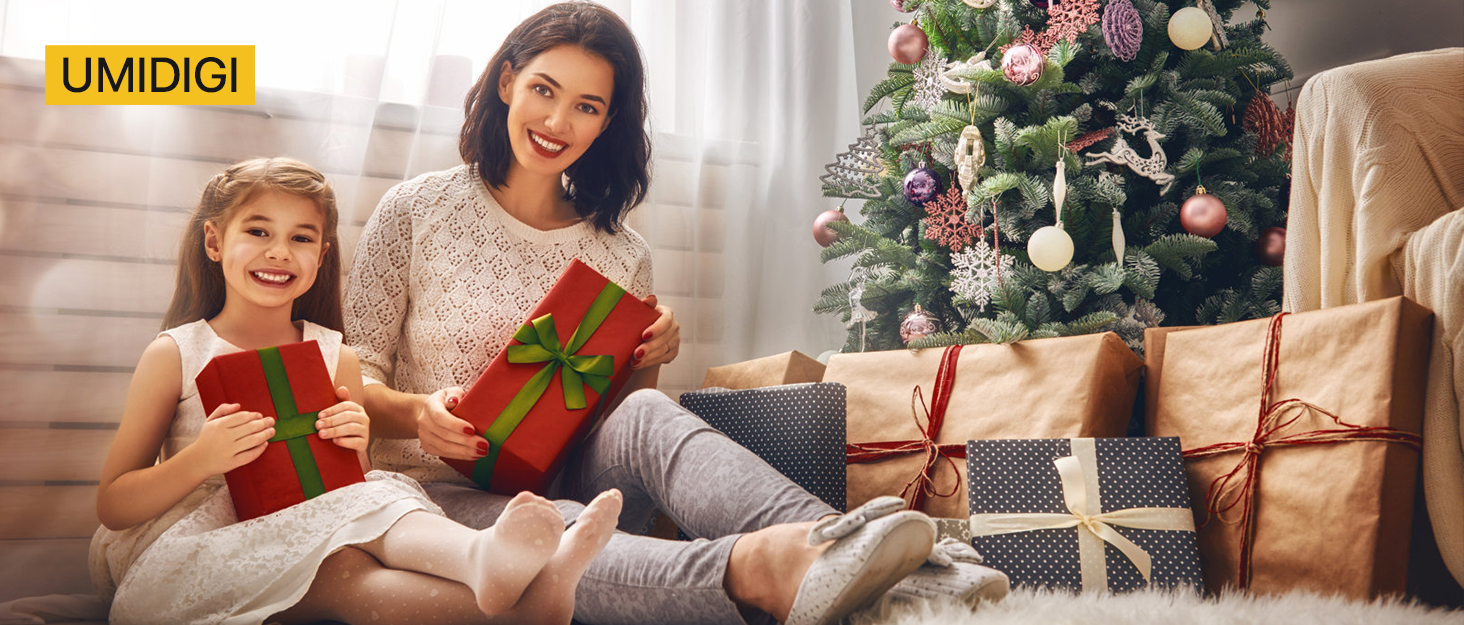 Decorated Christmas tree with wrapped gifts underneath. Two people sitting on the floor holding red and green wrapped presents, surrounded by additional gifts.